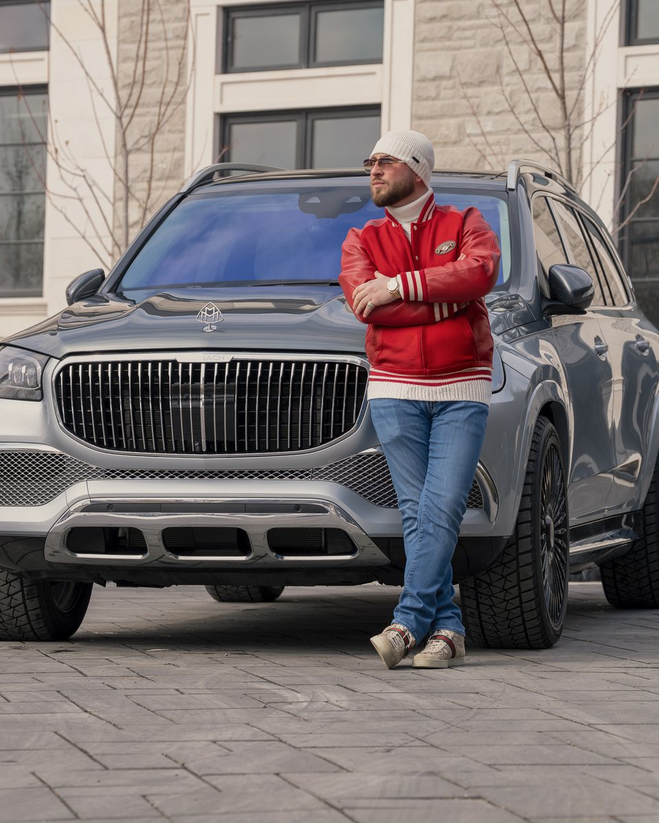 Man in a red jacket leaning against a silver Maybach SUV in front of a stone townhouse, luxury car portrait