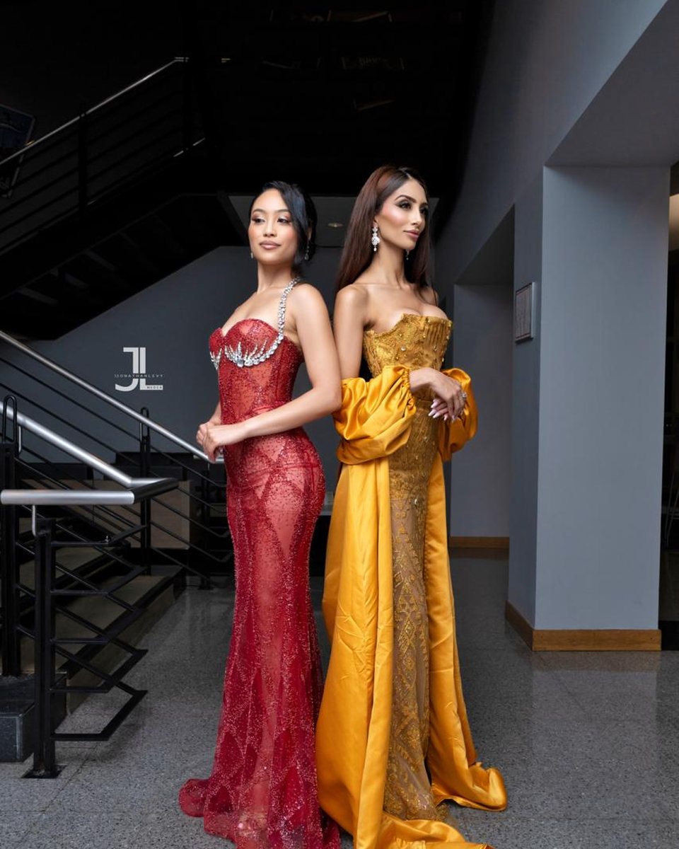 Two women in formal gowns, one in a red lace gown and one in a gold gown with a cape, standing together in a dark interior by a staircase